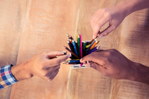 Bildet viser tre hender som tar hver sin fargeblyant fra en kopp full av fargeblyanter.