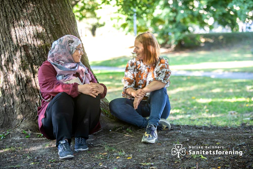 Bilde av Asmaa og Bente i samtale under et tre. Logoen til Norske Kvinners Sanitetskvinner