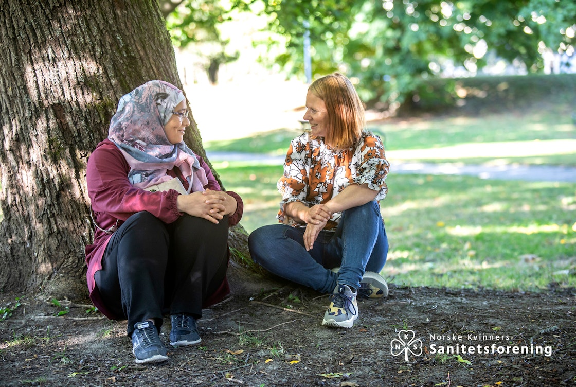 Bilde av Asmaa og Bente i samtale under et tre. Logoen til Norske Kvinners Sanitetskvinner