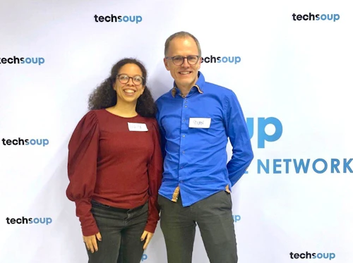 To personer står foran et banner med TechSoup logo på en konferanse og smiler vendt mot kamera.