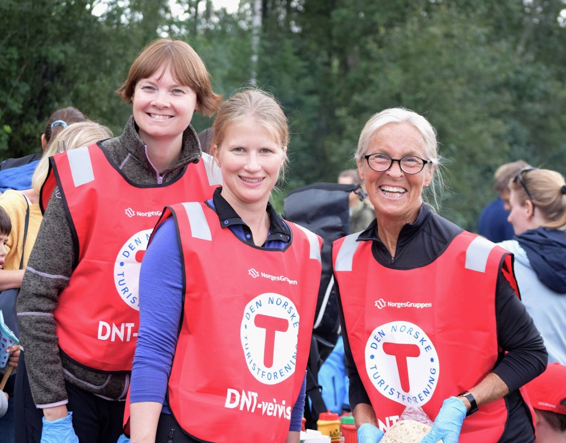 Tre kvinner i rød vest med logoen til Den Norske Turistforeningen smiler til kamera.