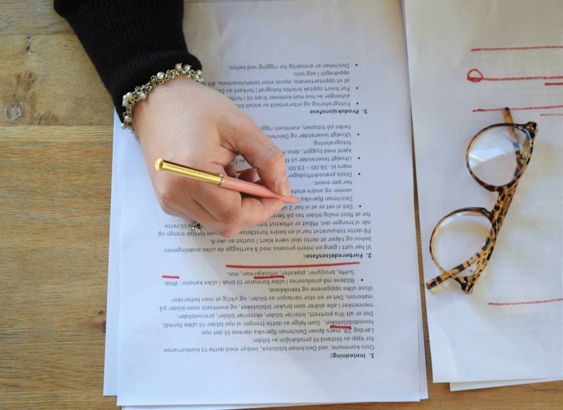 En håns skriver notater på et utprintet dokument. Et par briller hviler oppå dokumentet.
